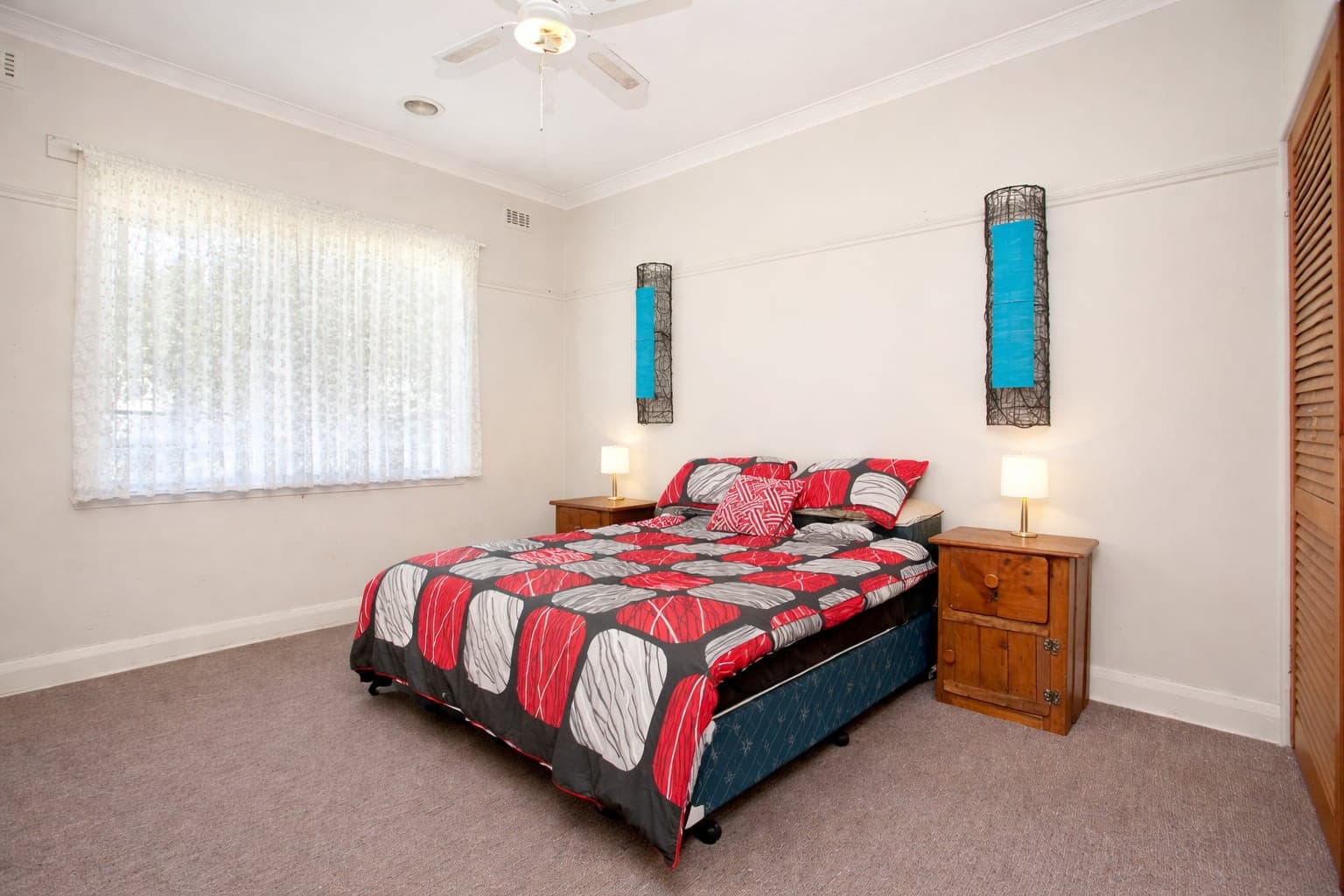 A carpeted bedroom with a large curtained window, a ceiling fan and a louvred built-in wardrobe.