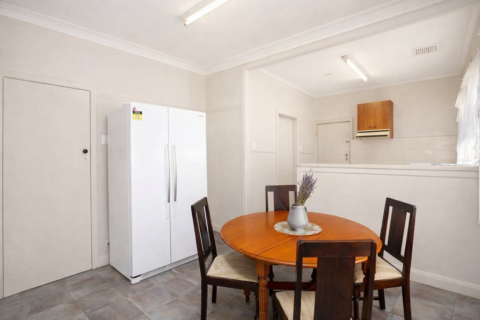 A tiled dining area with a round timber table and four chairs, a large fridge, and a serving opening through to the kitchen.