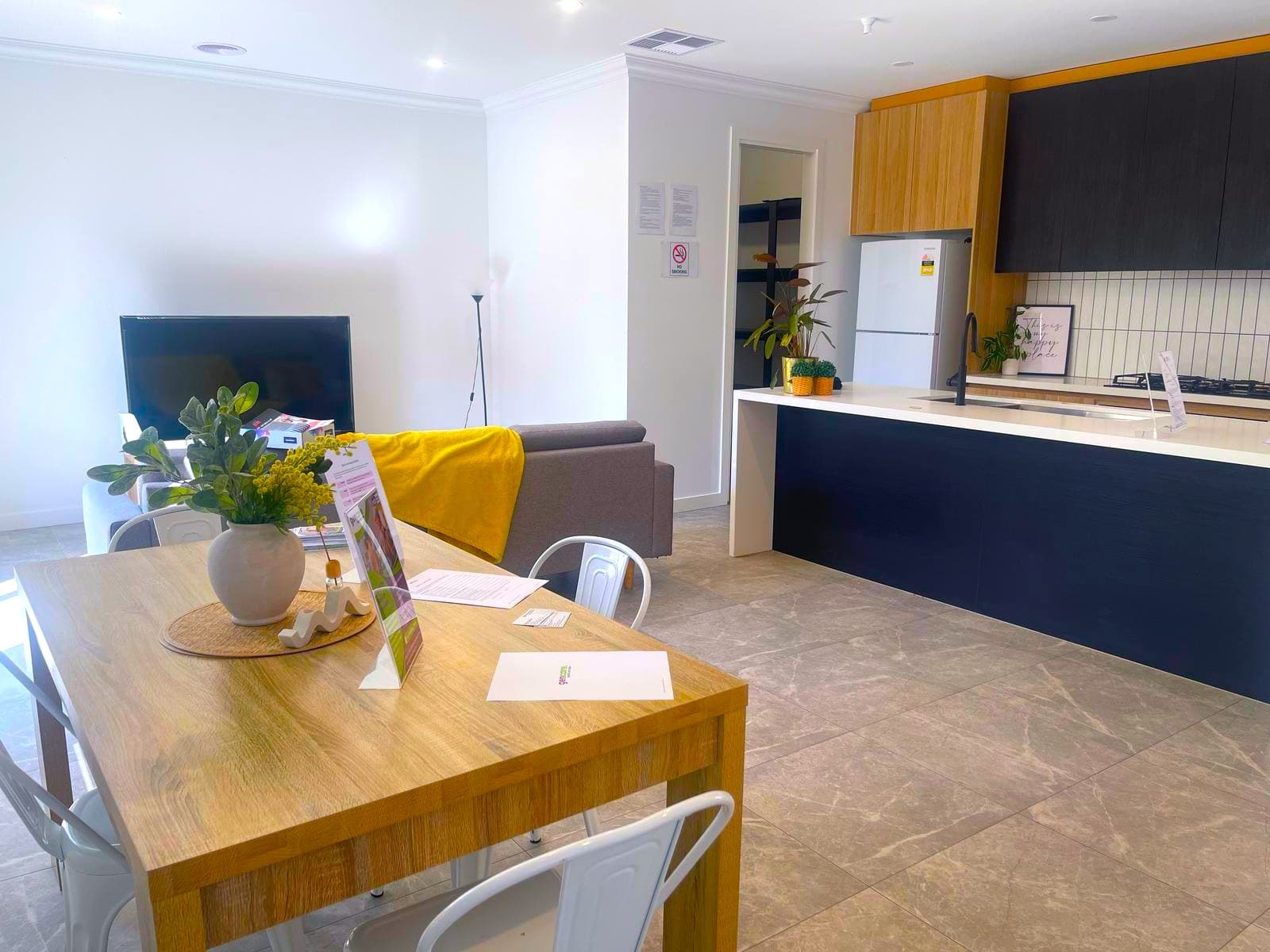 The open-plan living, dining and kitchen area of the Deanside home: a tiled floor running unbroken from a timber dining table, past a sofa, to a kitchen island, with a wide cased opening in the far wall.